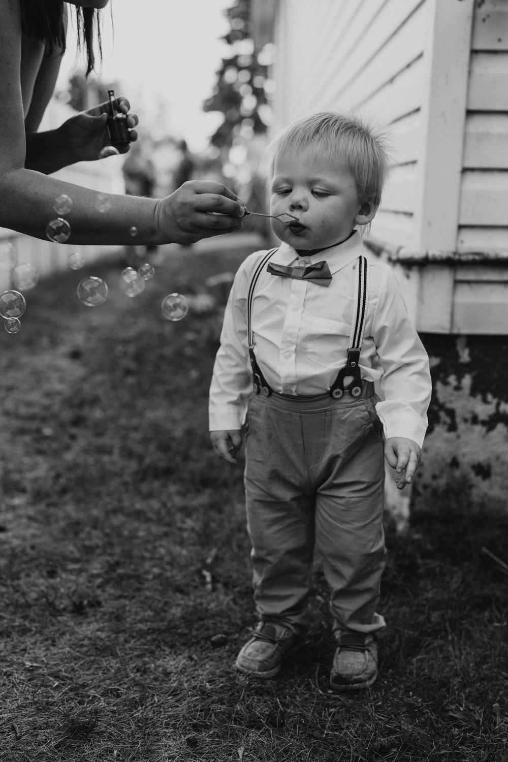 A toddler boy in suspenders and bow tie plays with bubbles outdoors at a wedding.