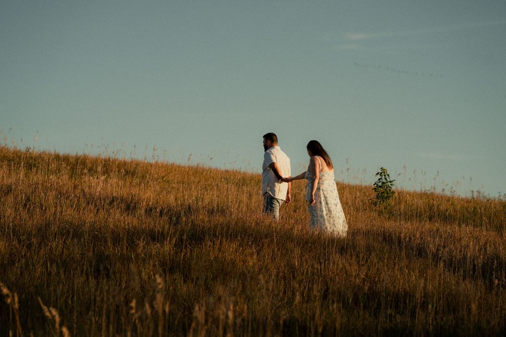 A couple holds hands while walking through a golden sunlit meadow under a clear sky.