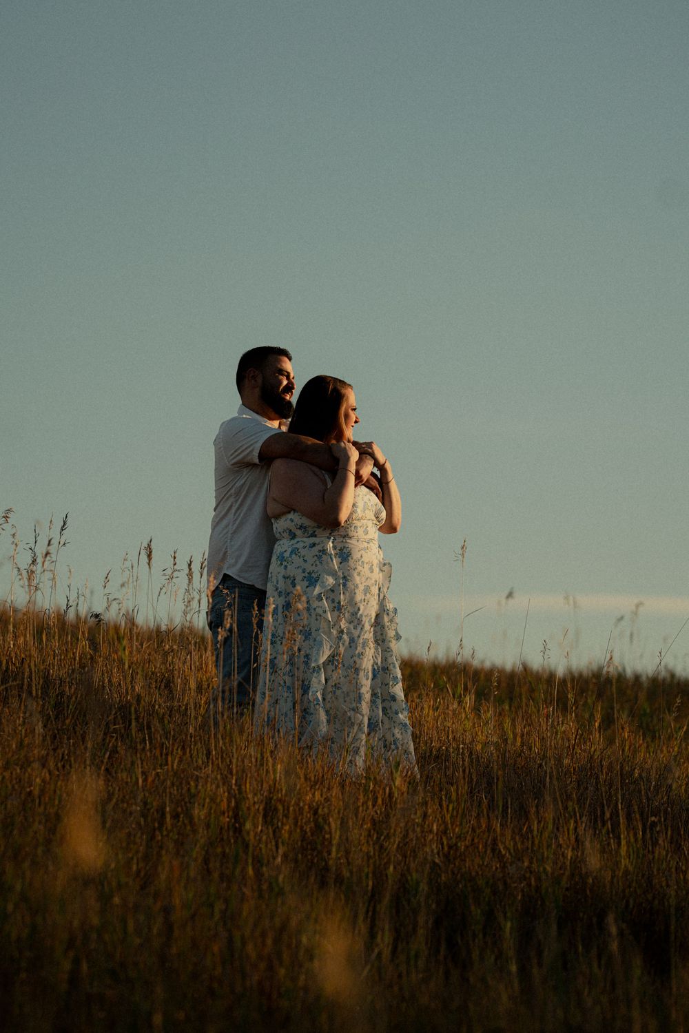 Couple embracing in golden field at sunset, woman in floral dress, romantic outdoor portrait photography.