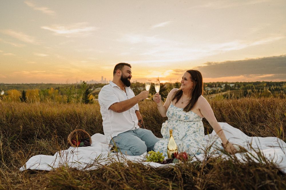 Couple toasting with wine glasses at a romantic sunset picnic in a golden field with grapes and basket.