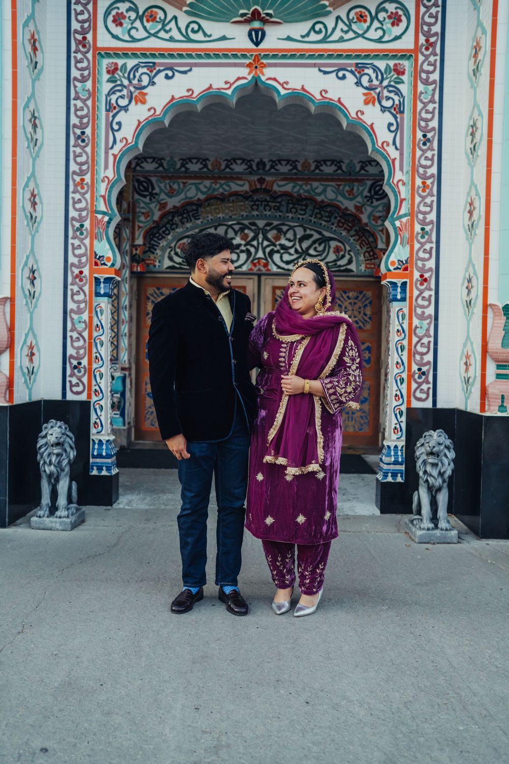 A couple in traditional South Asian attire poses before an ornately decorated temple entrance.
