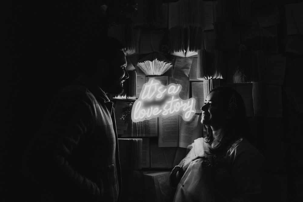 A couple stands in a dark room illuminated by a glowing neon sign on the wall.