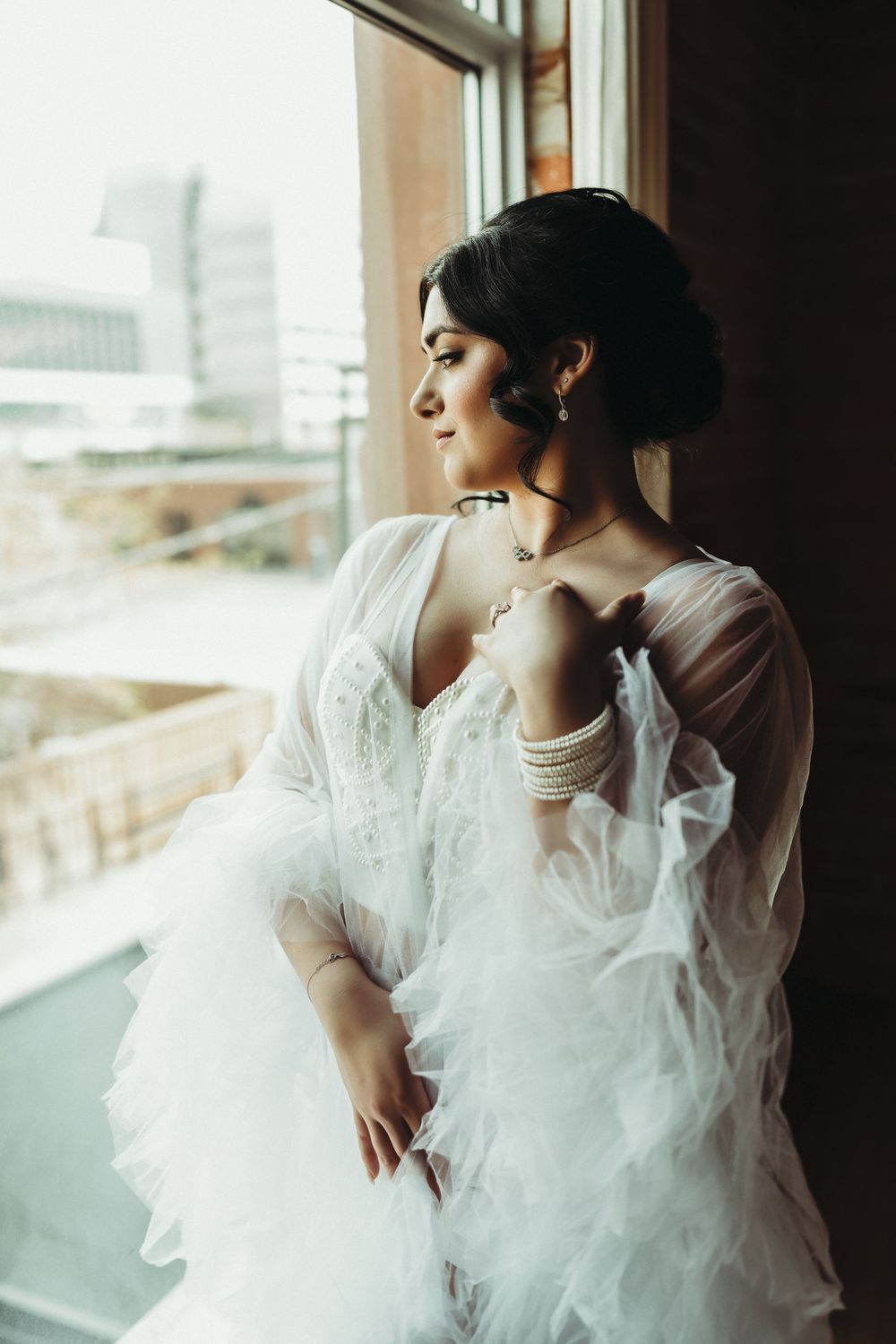 Elegant bride in a white ruffled gown standing by a window with city views, wearing gold jewelry.