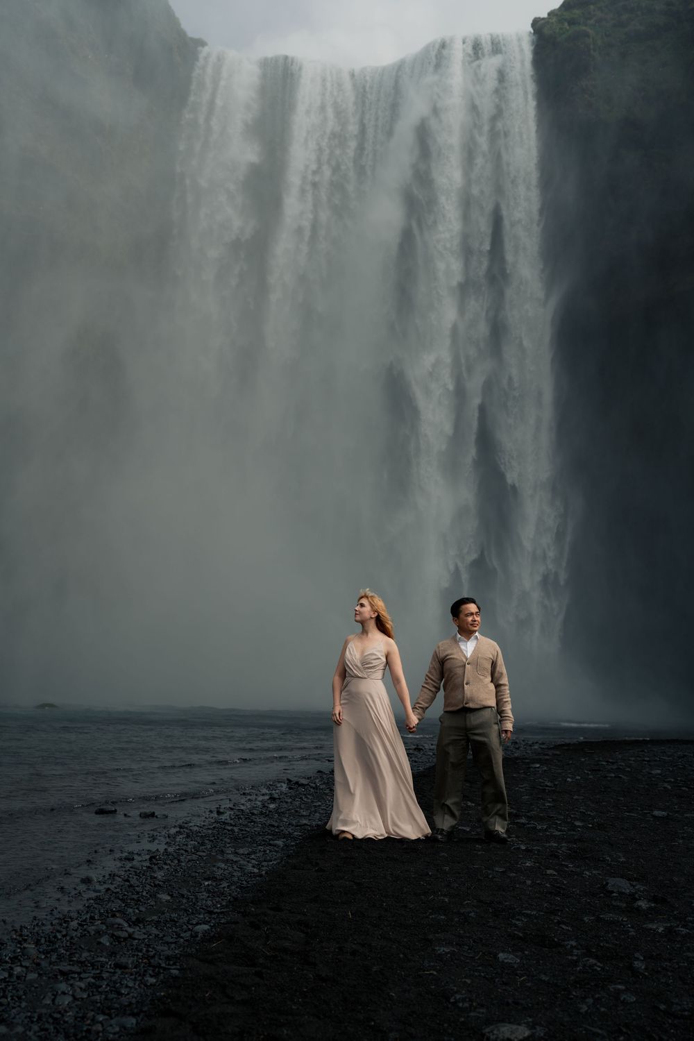 Couple holding hands in front of massive Skogafoss waterfall in Iceland, woman in flowing gown.