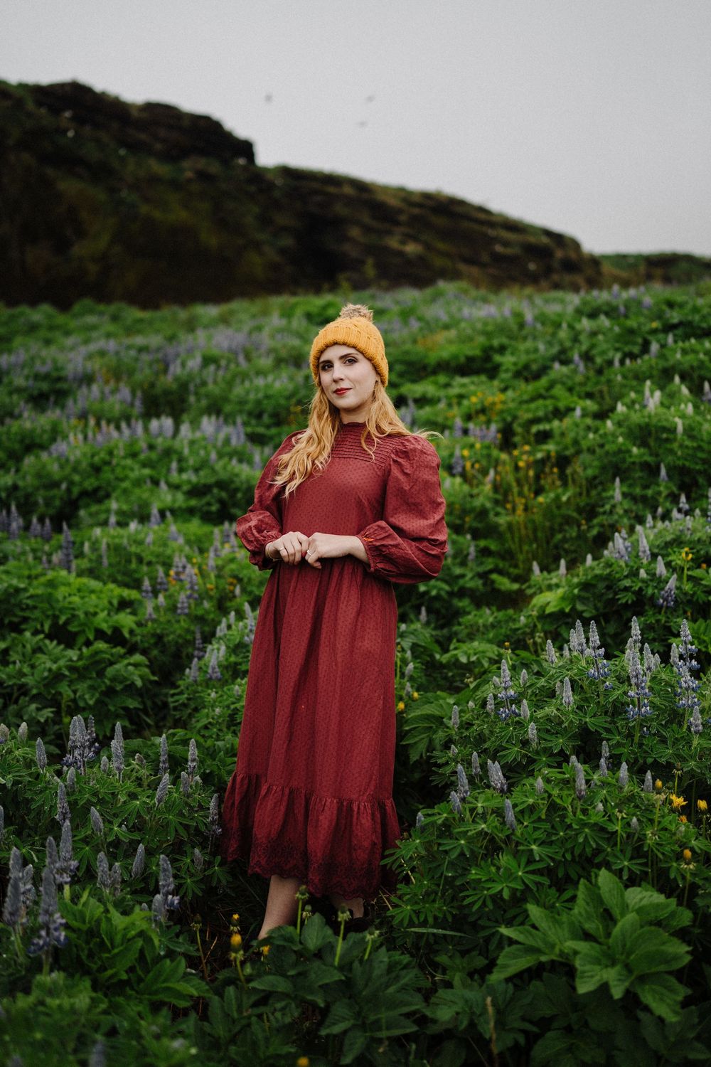 Woman in red dress and yellow beanie standing in lush green lupine flower field.