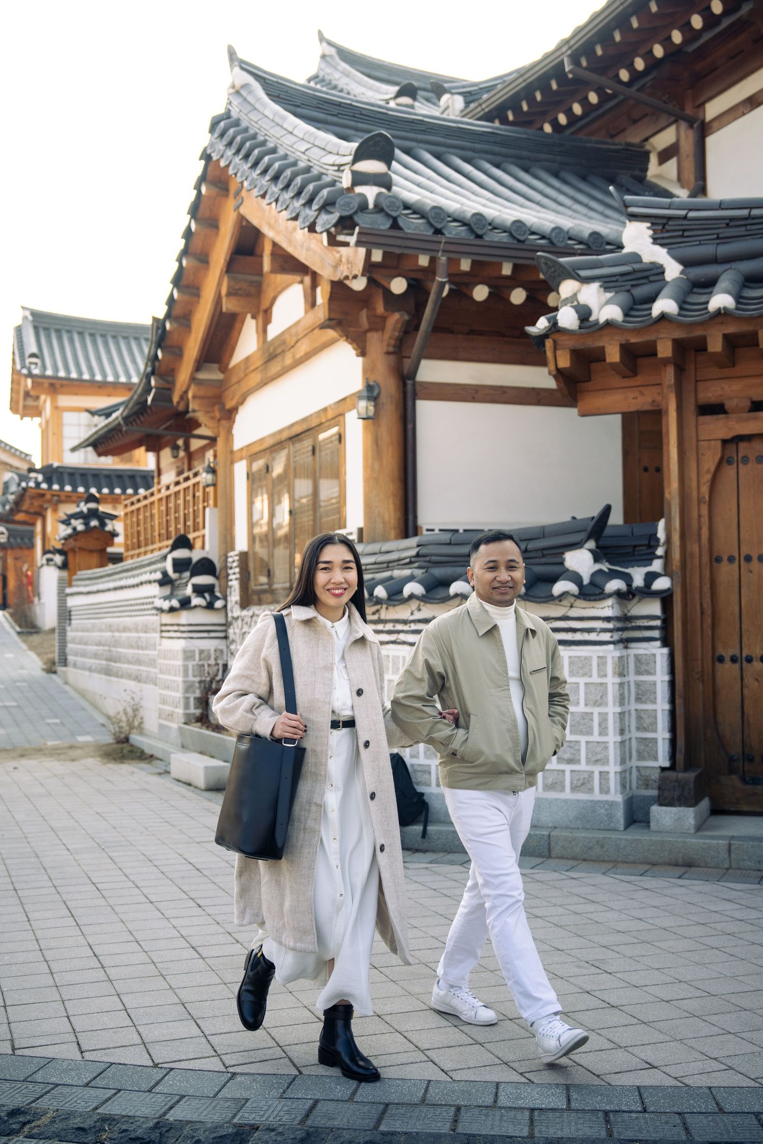 Two people in light colored outfits walk together along a traditional Korean hanok village pathway with tiled roofs.