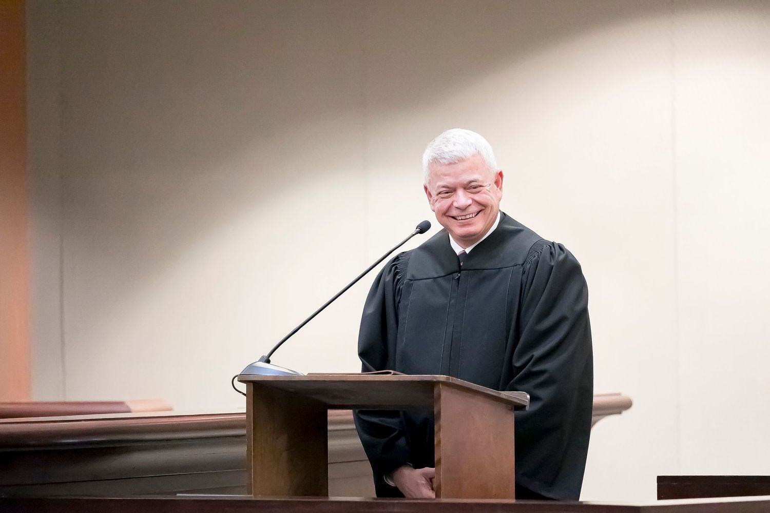 A person in a black judicial robe sits behind a wooden desk in a courtroom setting.