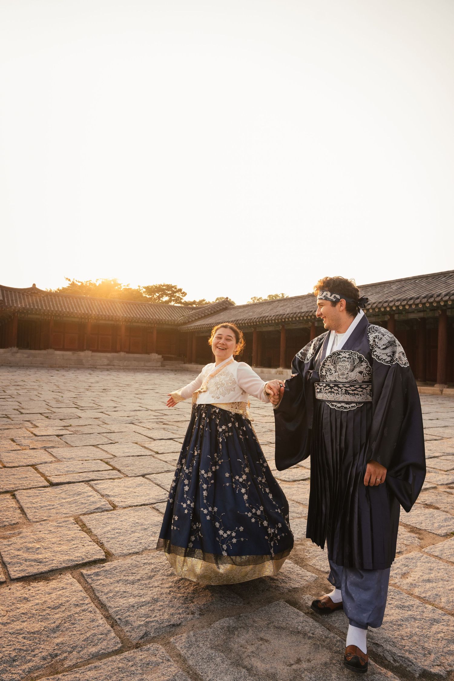 A couple in traditional Korean hanbok attire dances together at sunset on stone pavement.