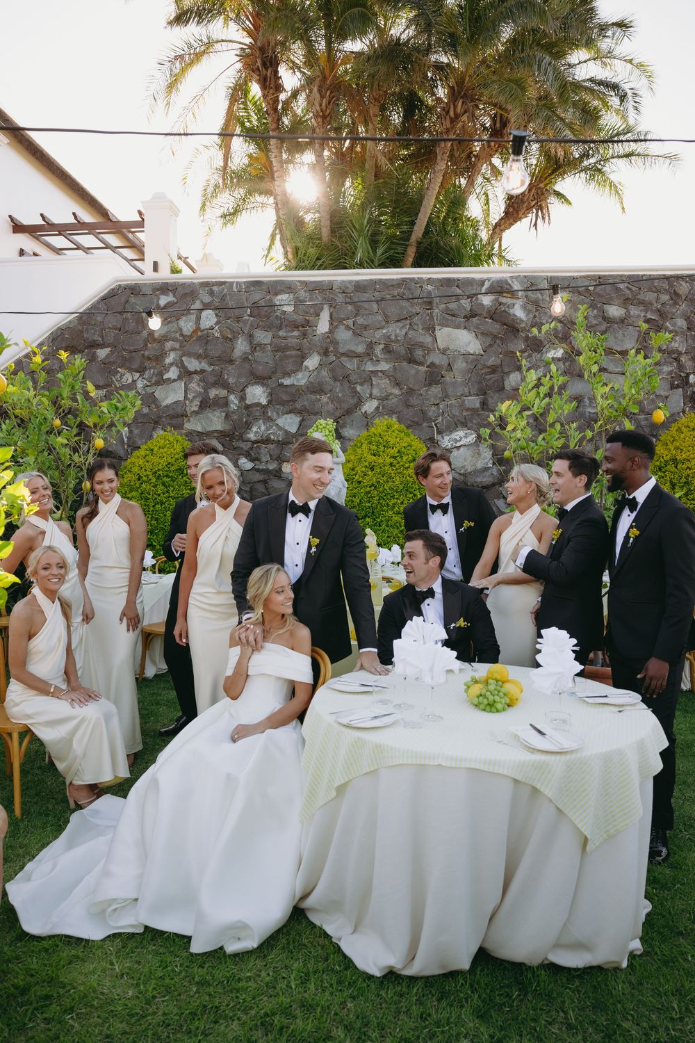 Wedding party members in formal attire seated on white chairs outdoors by a stone wall at sunset.
