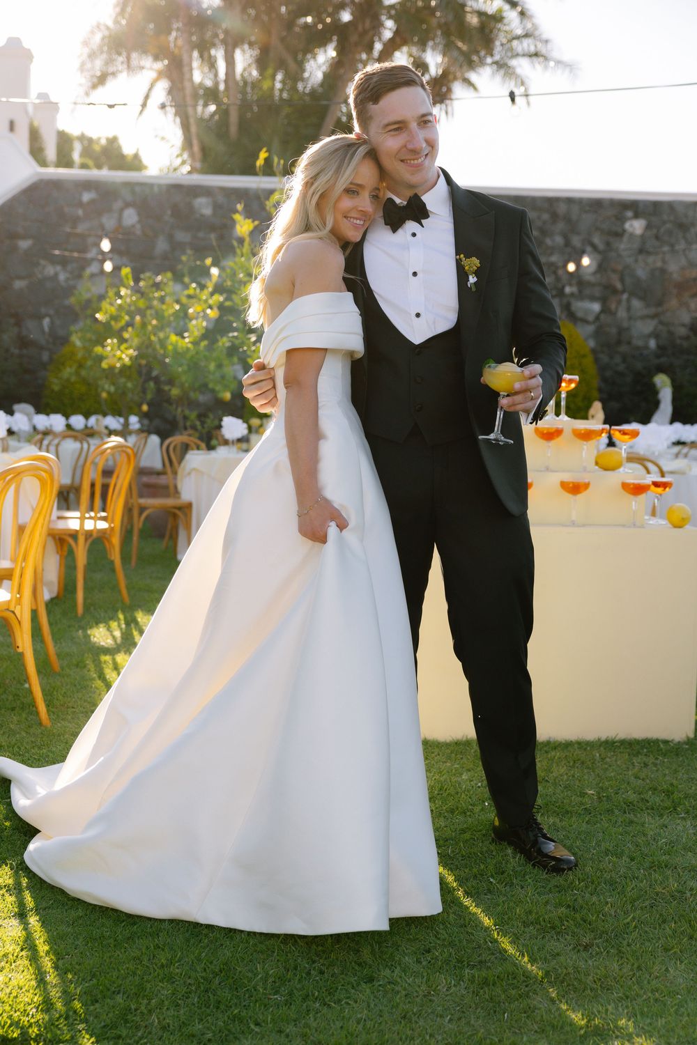 Elegant wedding couple dances outdoors at sunset with golden light filtering through trees in garden reception.