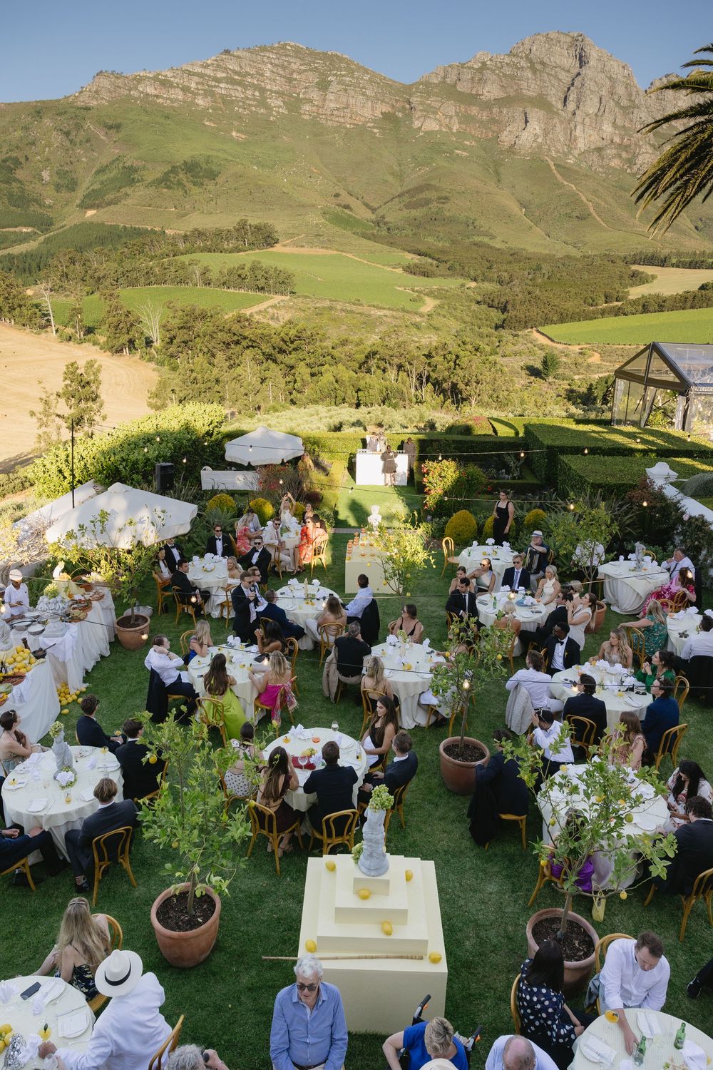 Outdoor garden wedding reception with white-clothed tables and guests seated under mountain views and natural scenery.