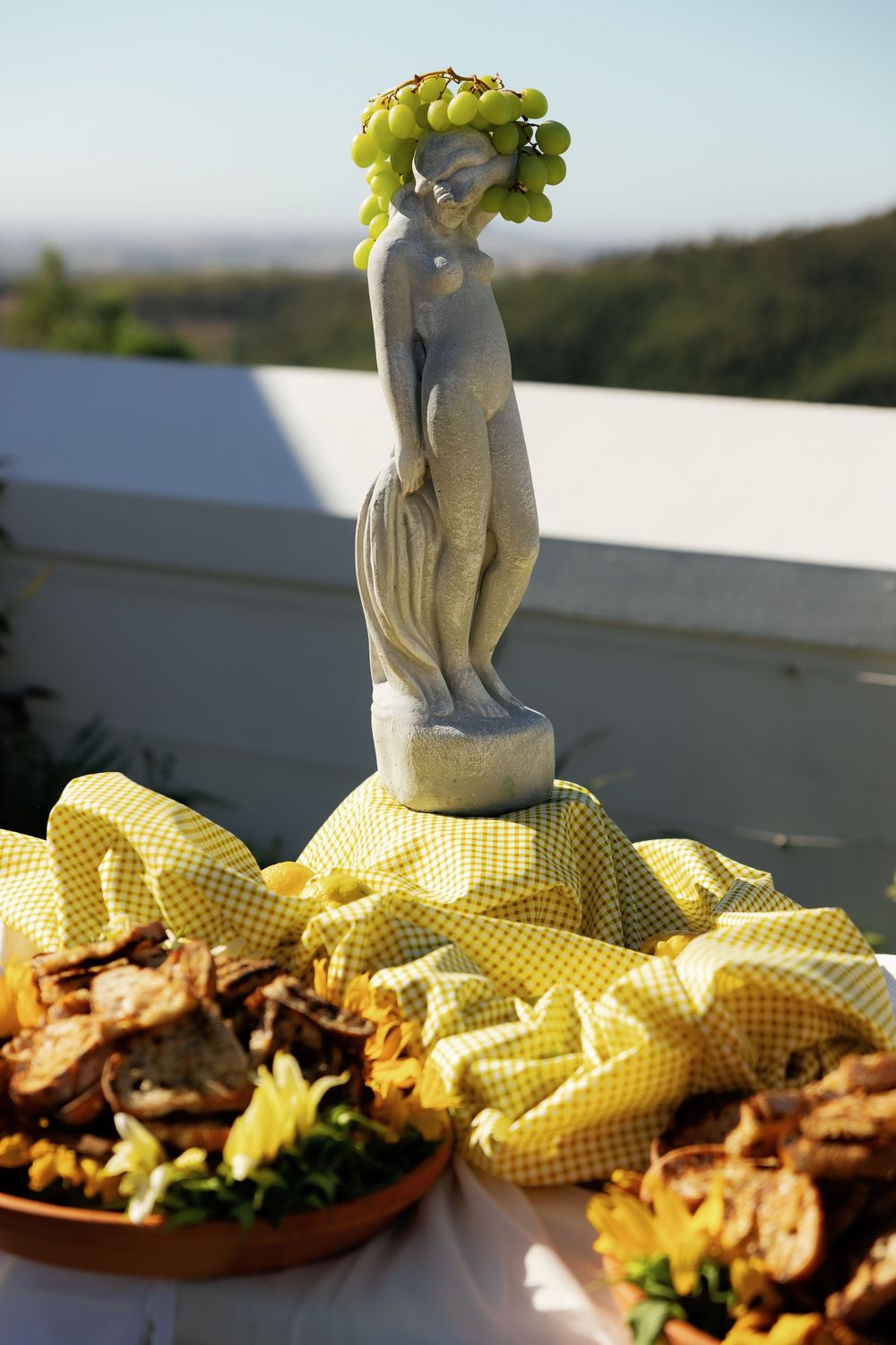 Stone cherub statue sits atop yellow gingham tablecloth with appetizers at outdoor event.
