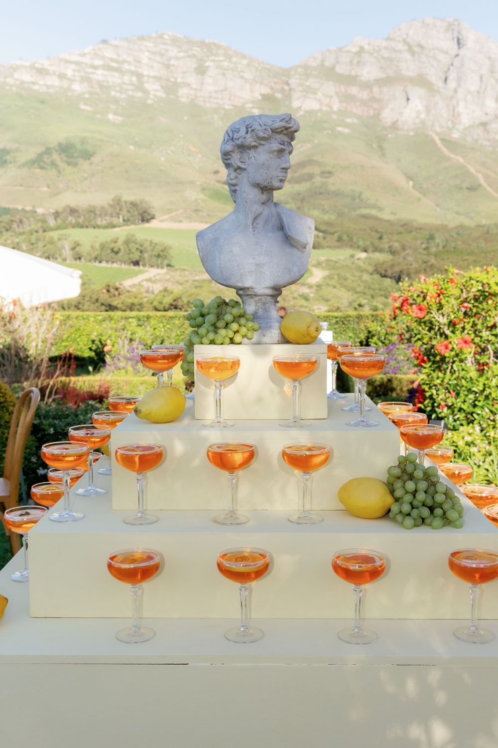 Elegant cocktail display with orange drinks arranged in tiers beneath a classical bust statue against mountain backdrop.
