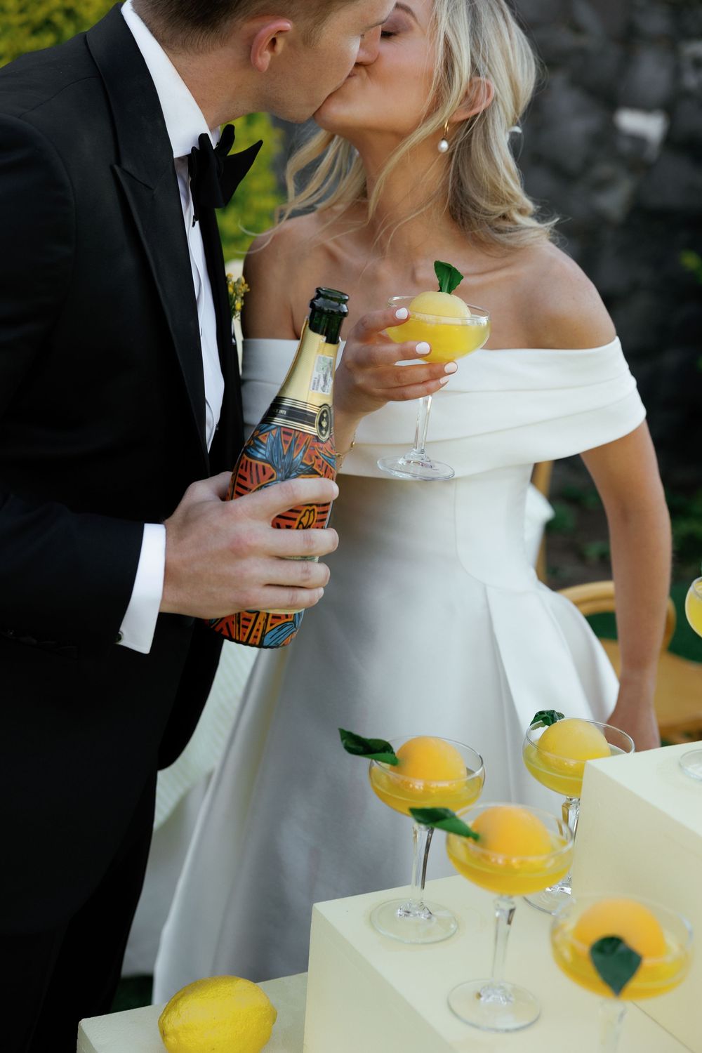A couple in formal wedding attire shares yellow cocktails at a reception.