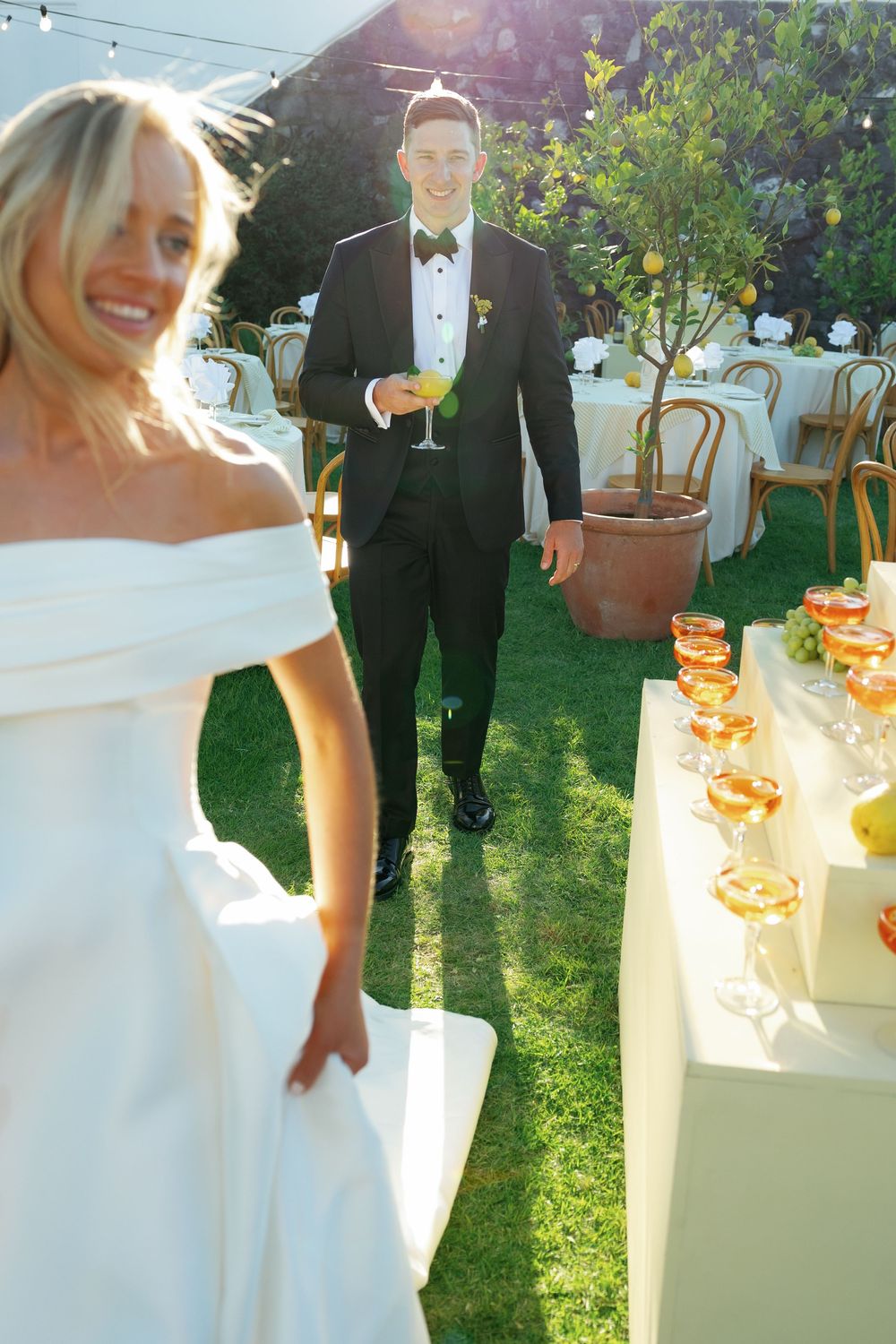 A couple in wedding attire holding champagne glasses stands in a sunlit garden during an outdoor celebration.