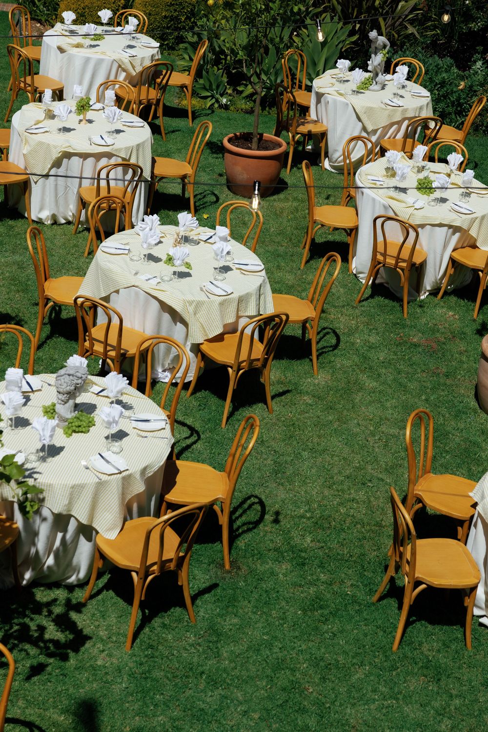 Elegant outdoor wedding reception setup with round tables covered in white linens and wooden bentwood chairs on a lawn.