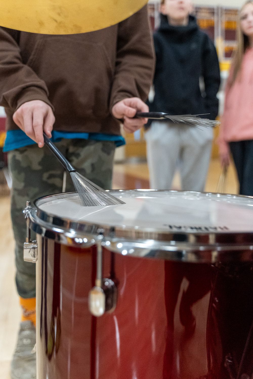 Young student playing a red bass drum with wire brushes during a music class or band practice.