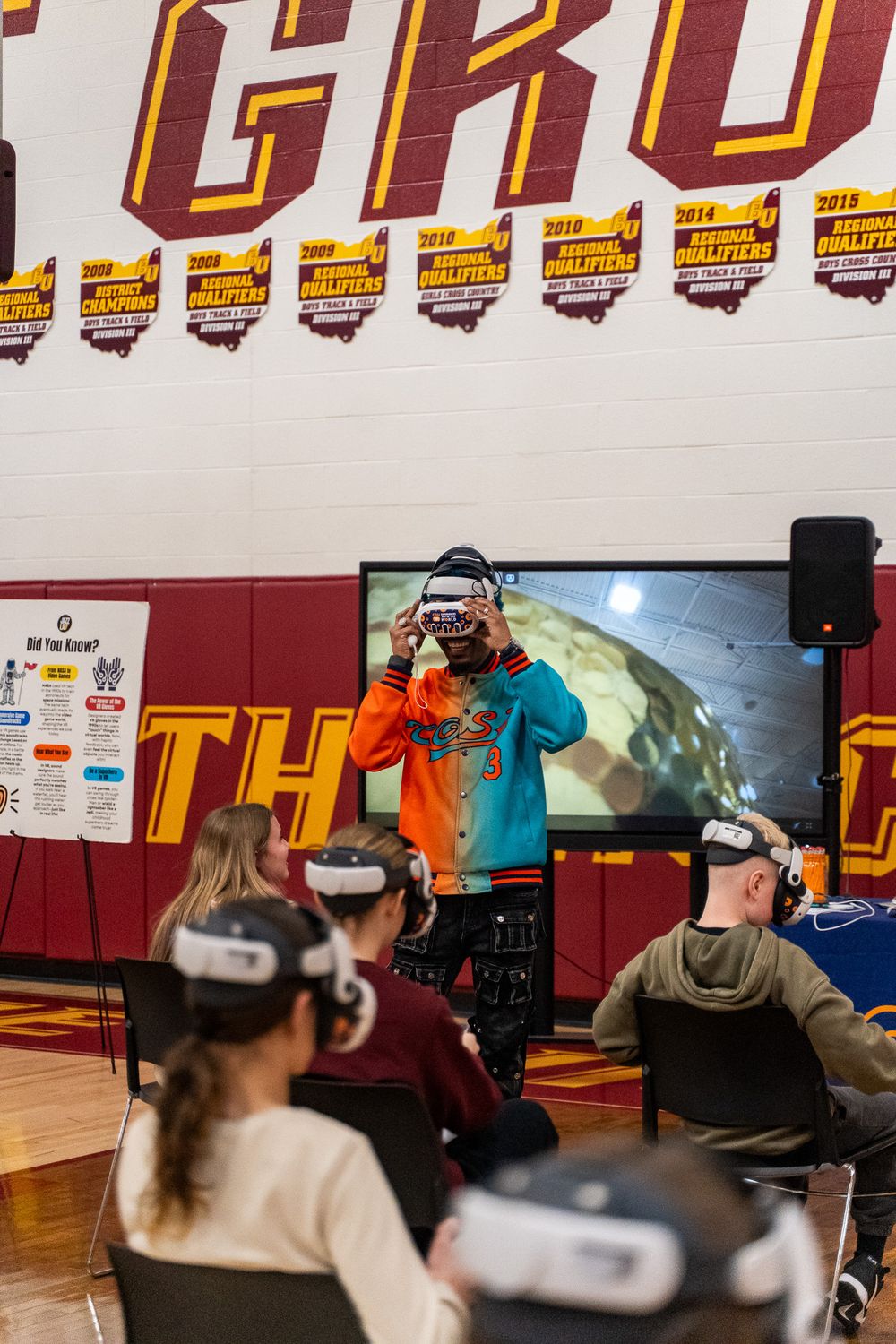 Students wearing VR headsets in a school gym while an instructor demonstrates virtual reality technology on a large screen.