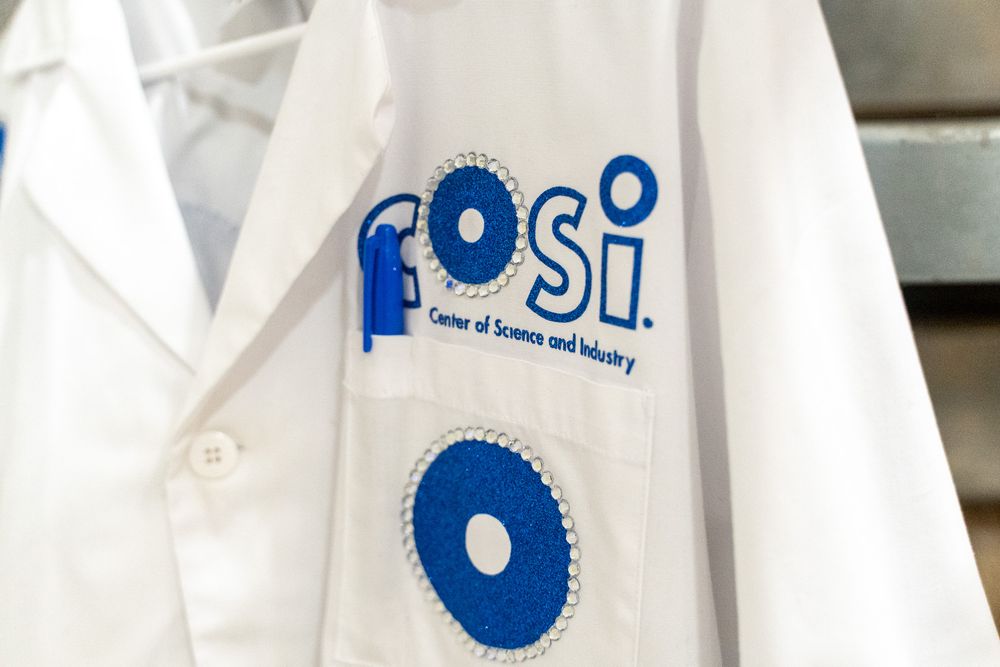 White lab coat with COSI Center of Science and Industry logo in blue, hanging on display.