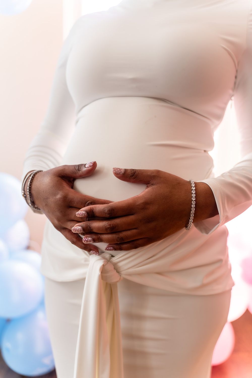 Pregnant woman in white dress cradling baby bump with hands adorned with bracelets at a gender reveal party.