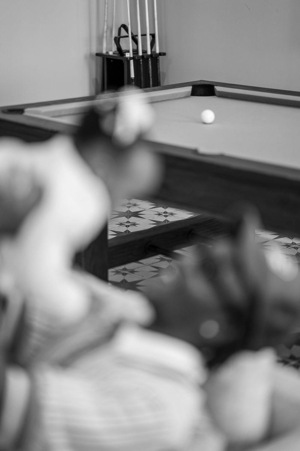 Black and white photo of a pool table with cue ball and billiard cues in rack, blurred figures in foreground.