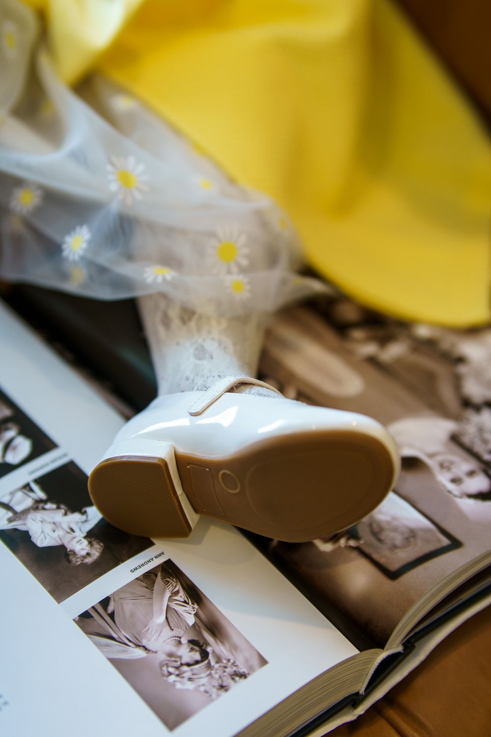 White patent leather baby shoes on open photo album with yellow hat and daisy tulle fabric.