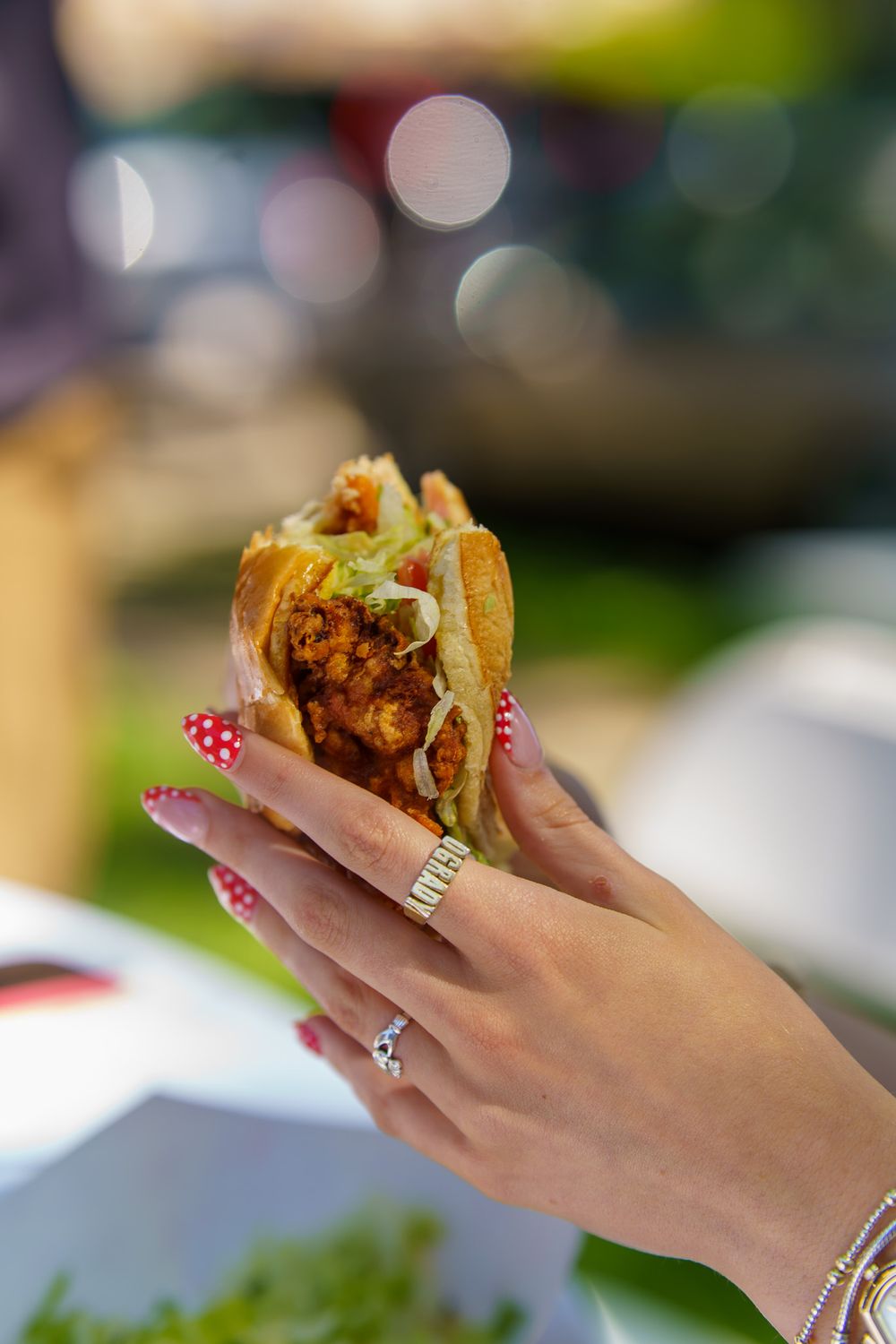 A woman with colorful nails and rings holds a crispy fried chicken taco outdoors.