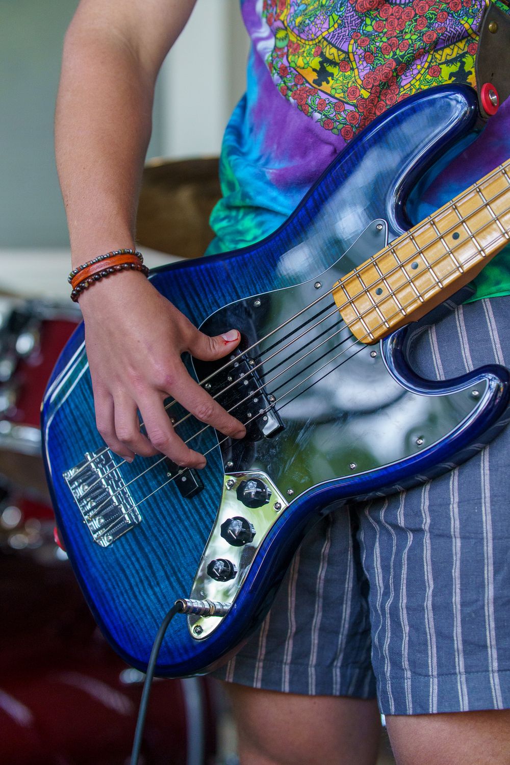 Musician playing a blue flame maple Fender Jazz Bass guitar while wearing a tie-dye shirt and gray shorts.