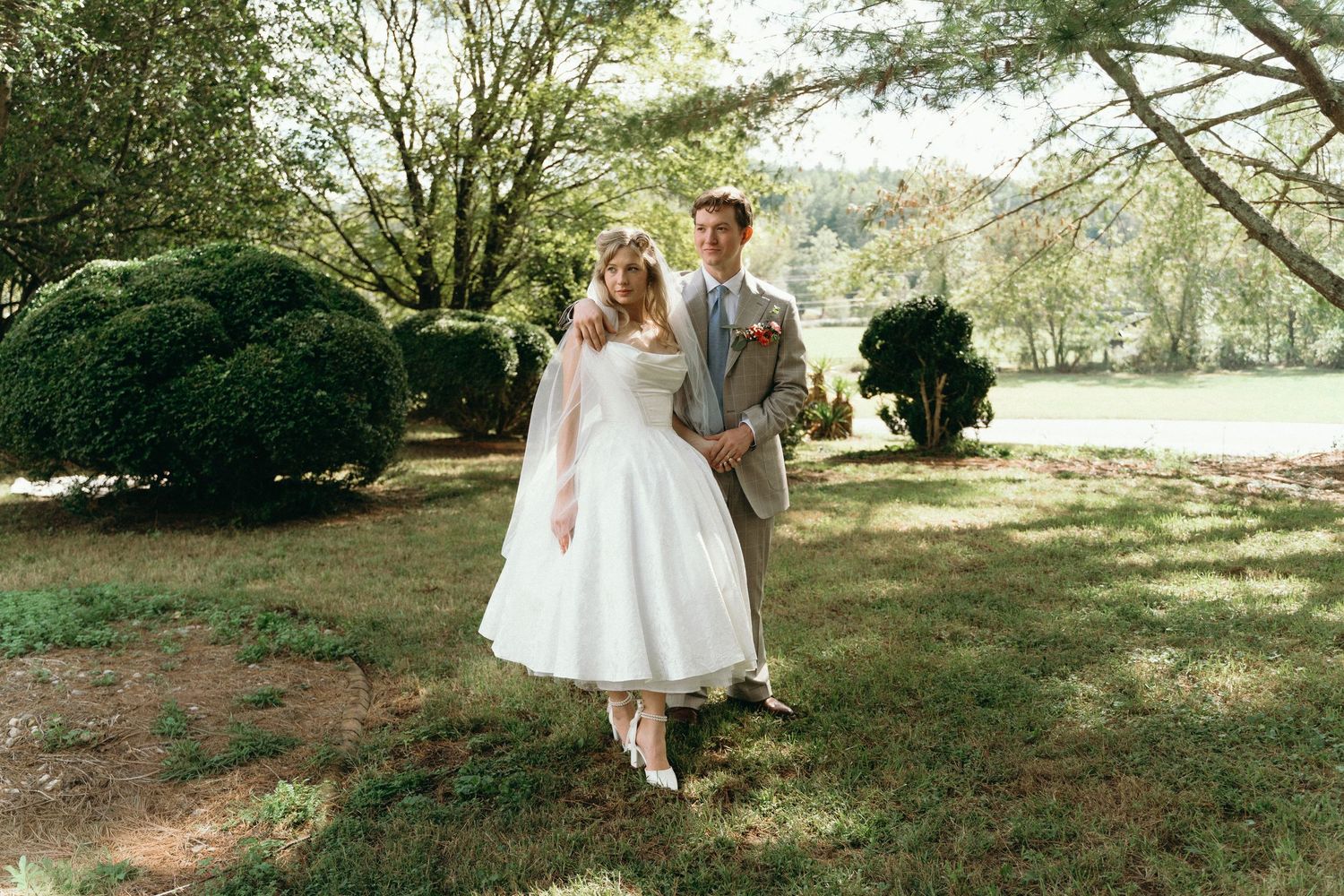 A couple in wedding attire walks hand in hand through a sunlit garden with lush trees and manicured shrubs. East Coast Luxury wedding photos