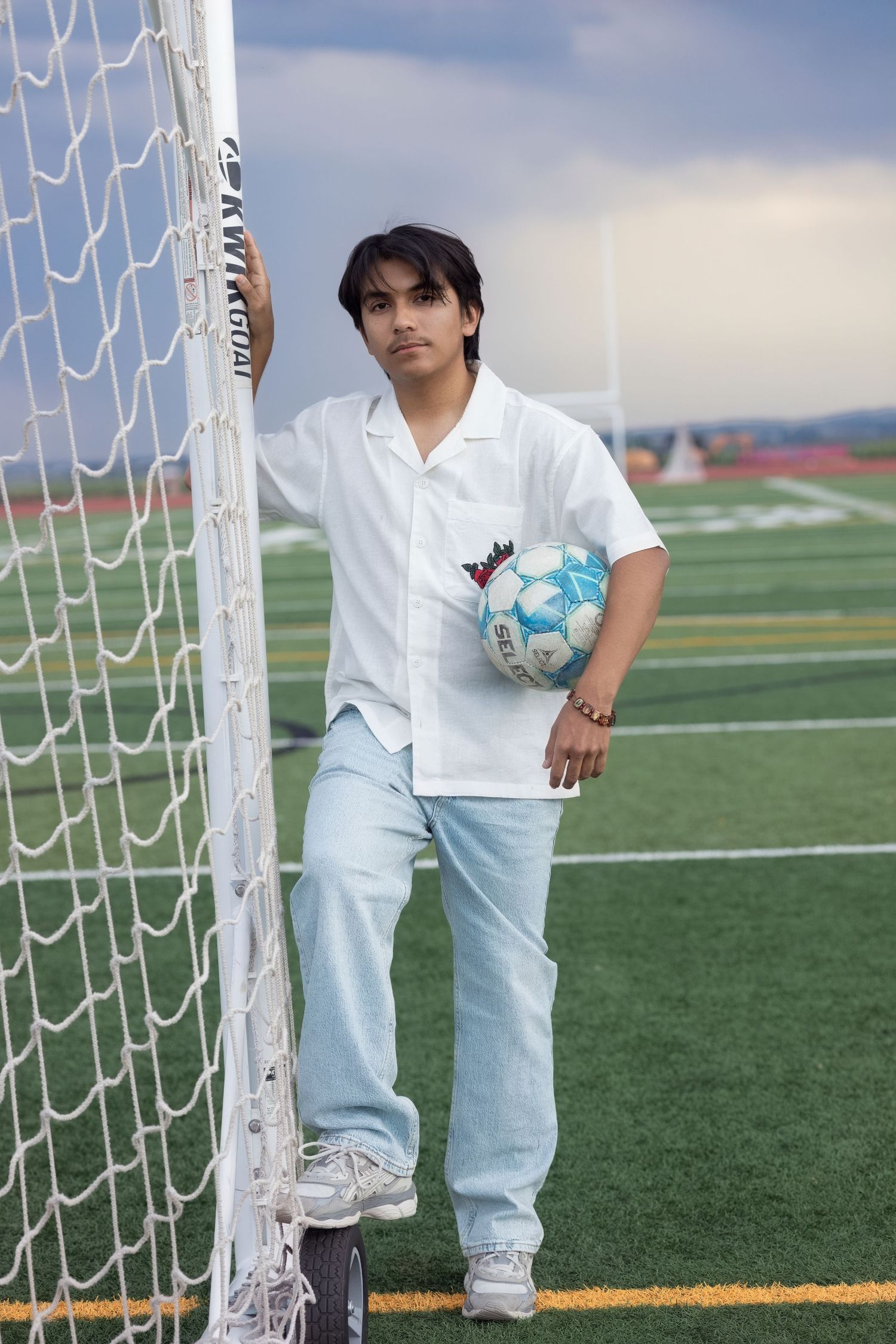 Figure in white polo shirt and light jeans stands by soccer goal on artificial turf field holding blue soccer ball.