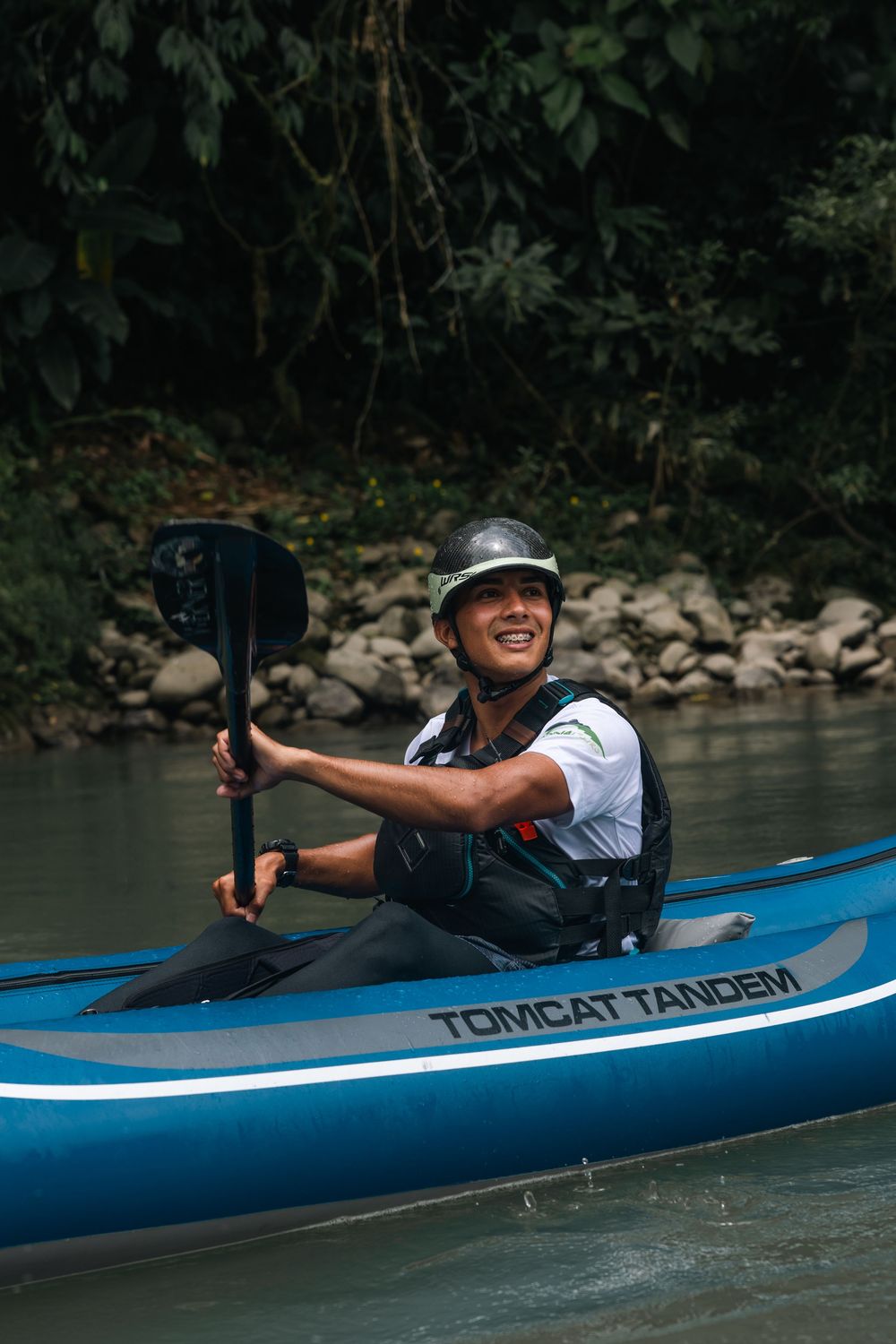 A kayaker in a blue Tomcat vessel paddles through calm river waters with forested banks.