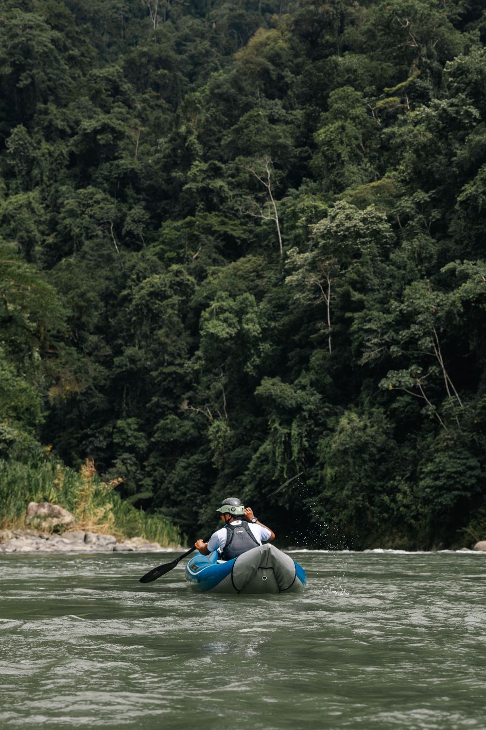 A blue raft floats down a wide river surrounded by dense green forest slopes.