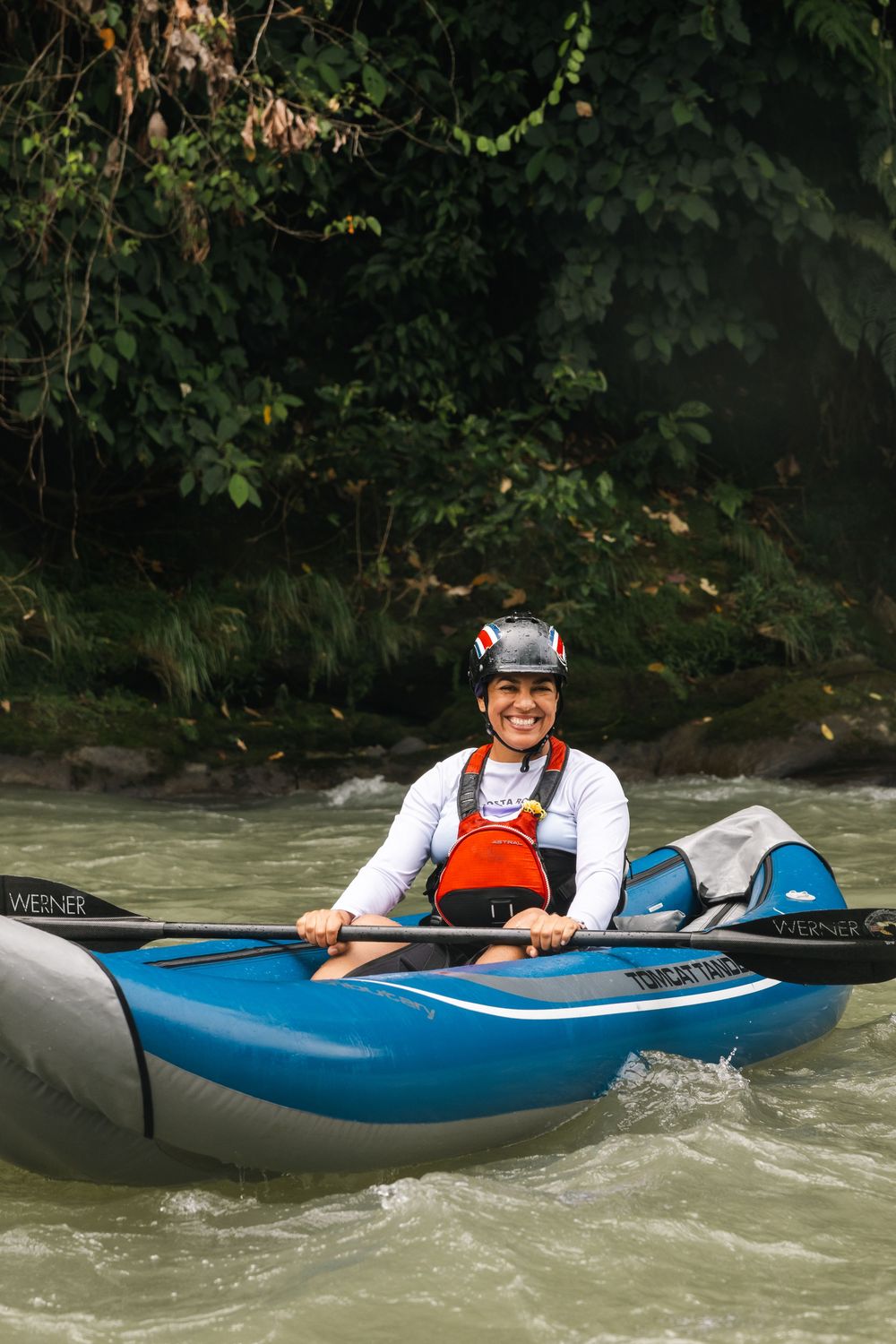 Kayaker in blue boat smiles while navigating river rapids.
