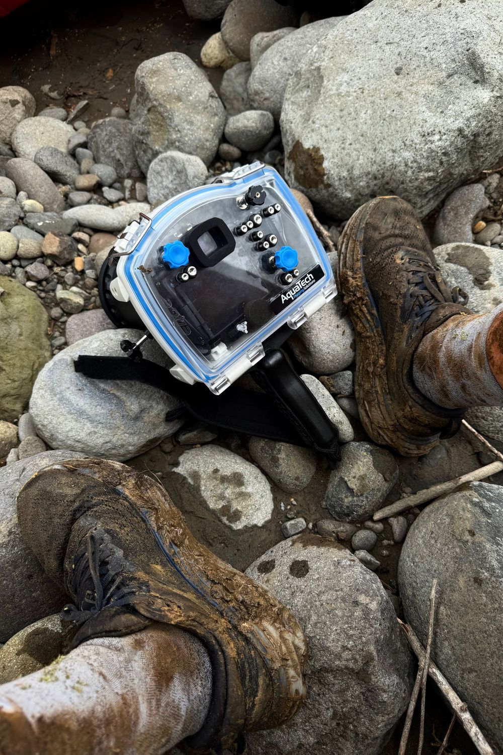 Blue camera equipment positioned among rocky terrain and driftwood on a beach or riverbed.