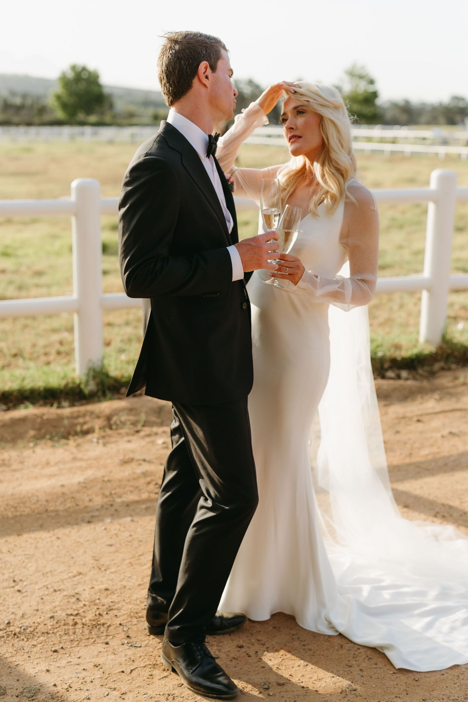 A couple in formal wedding attire shares a tender moment outdoors on a sunny day near a white fence.