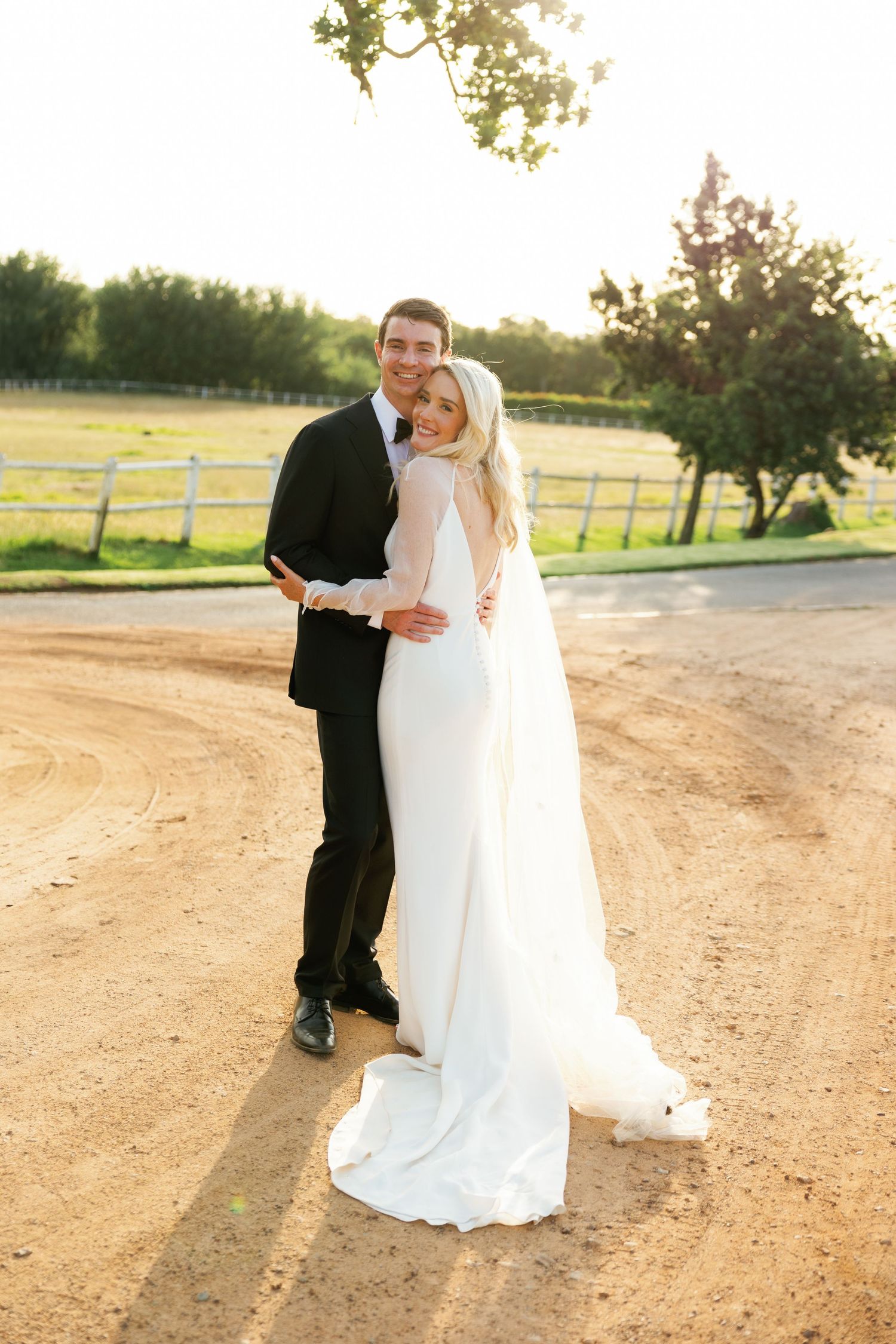 A couple in wedding attire embrace on a dirt path with a rustic fence and countryside setting at sunset.