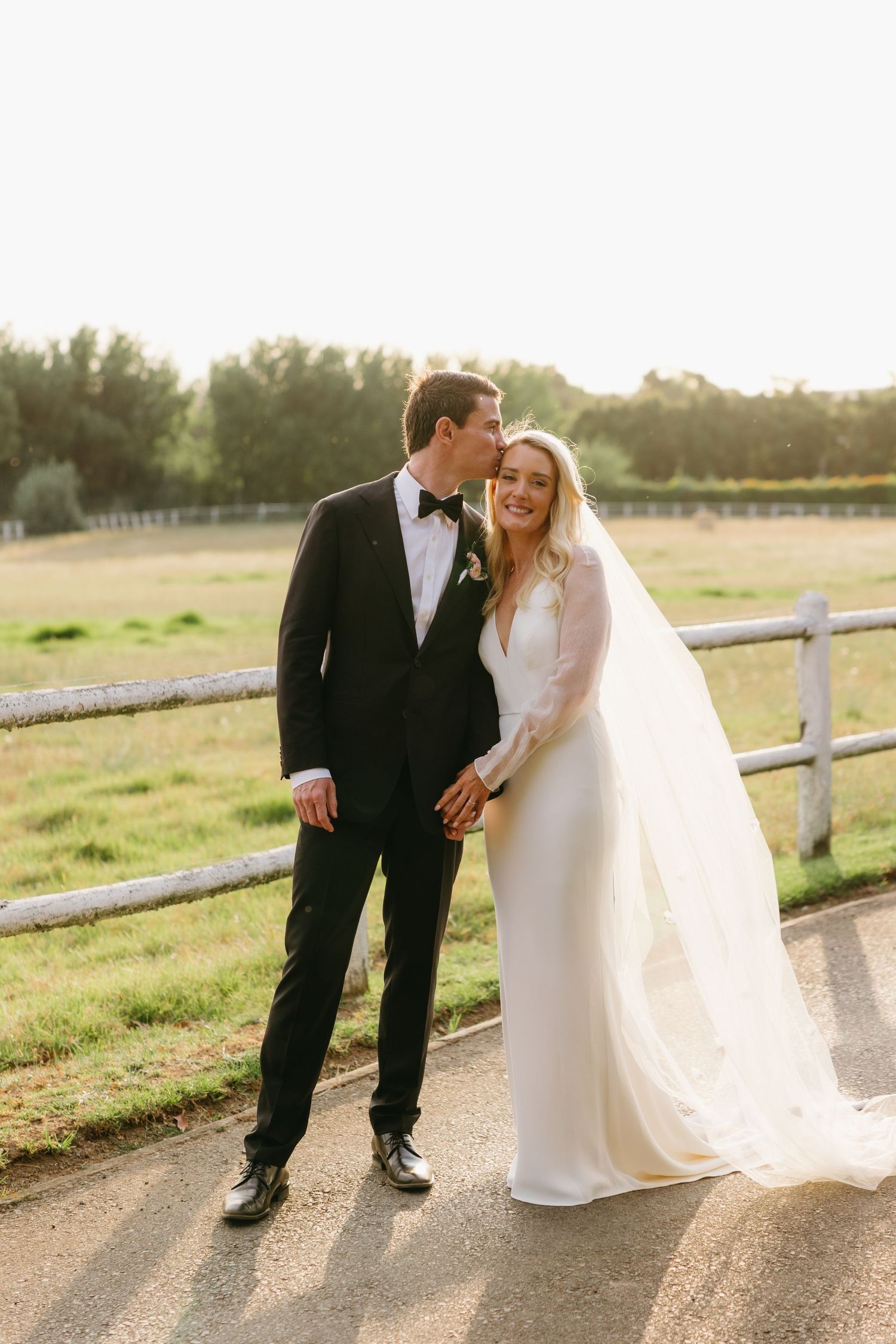 A newlywed couple shares a joyful moment together on a country path with white fencing at golden hour.