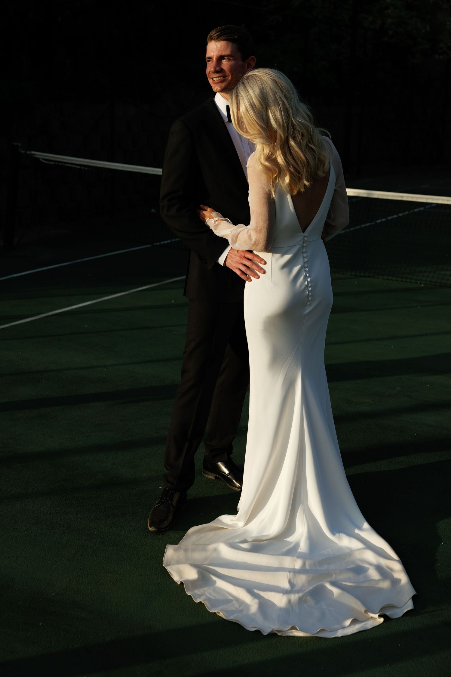 A couple dances together on a tennis court at dusk, with the bride wearing a flowing white wedding dress.