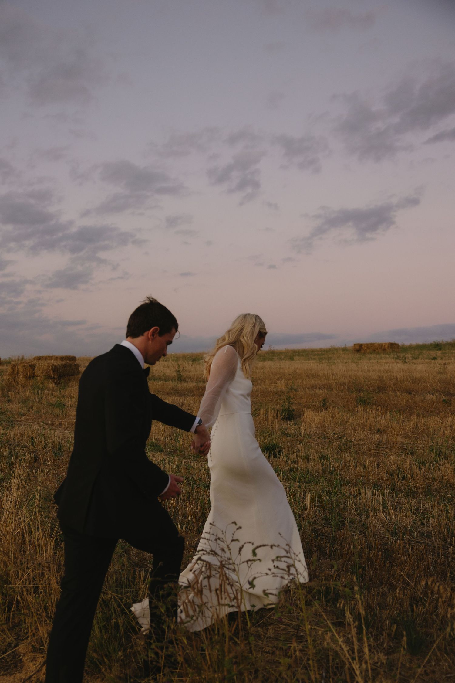 A couple in formal attire walks through a golden field at dusk with dramatic cloudy sky.