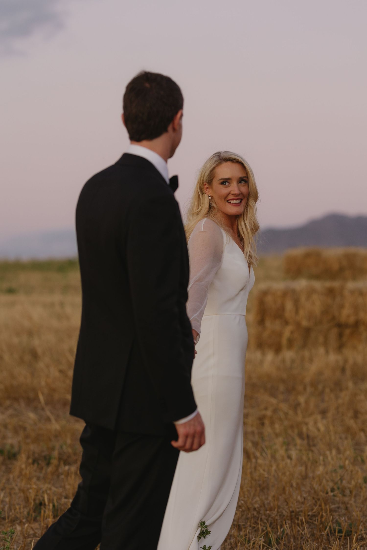 A couple shares a candid moment in formal attire against a dusky rural landscape with mountains.