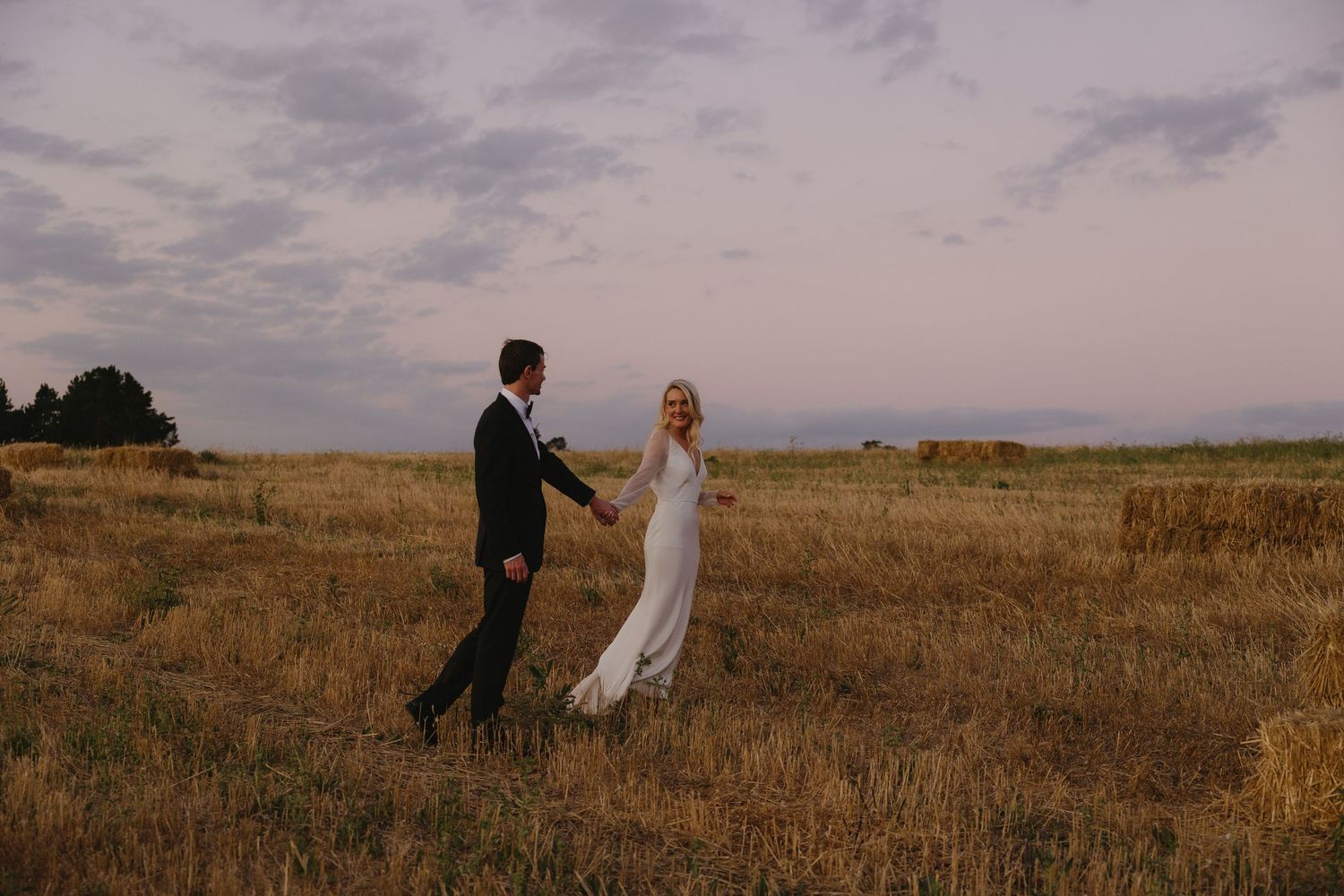 Newlyweds stand together in a golden field at dusk with mountains visible in the distance.
