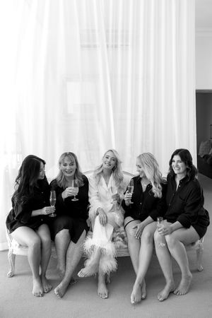 A group of women in black leotards sit in a row against a white wall with their legs outstretched.