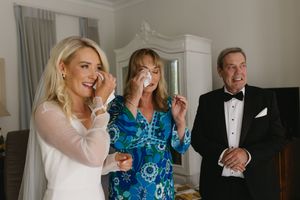 Three people in formal attire share an emotional moment at an indoor gathering, with two wiping tears and one smiling.
