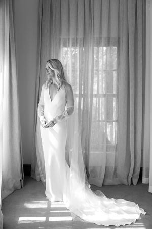 Black and white photograph of flowing wedding dress silhouette against sheer curtained windows with natural light.