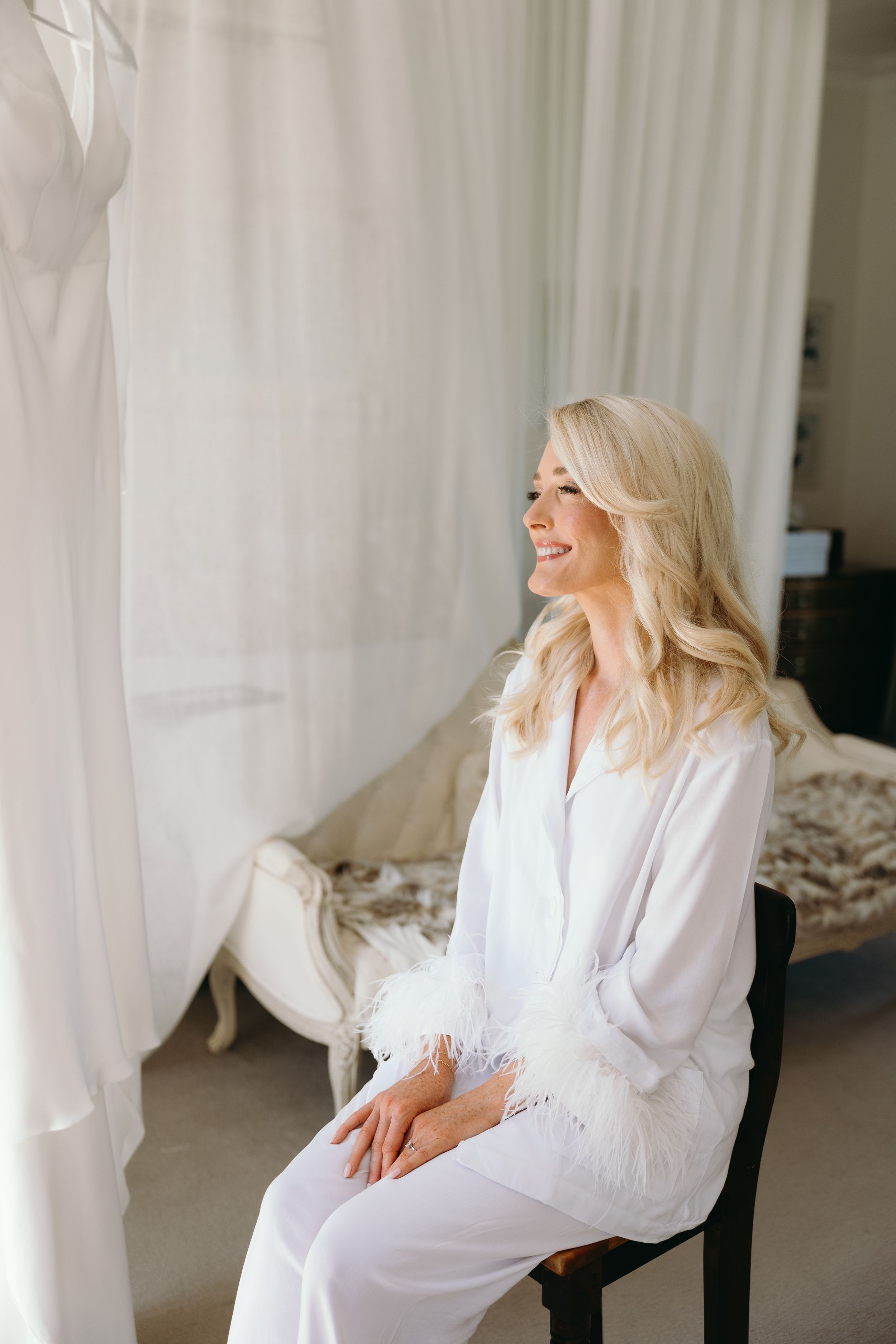A person in a white feather-trimmed robe sits smiling by a window during wedding preparations.