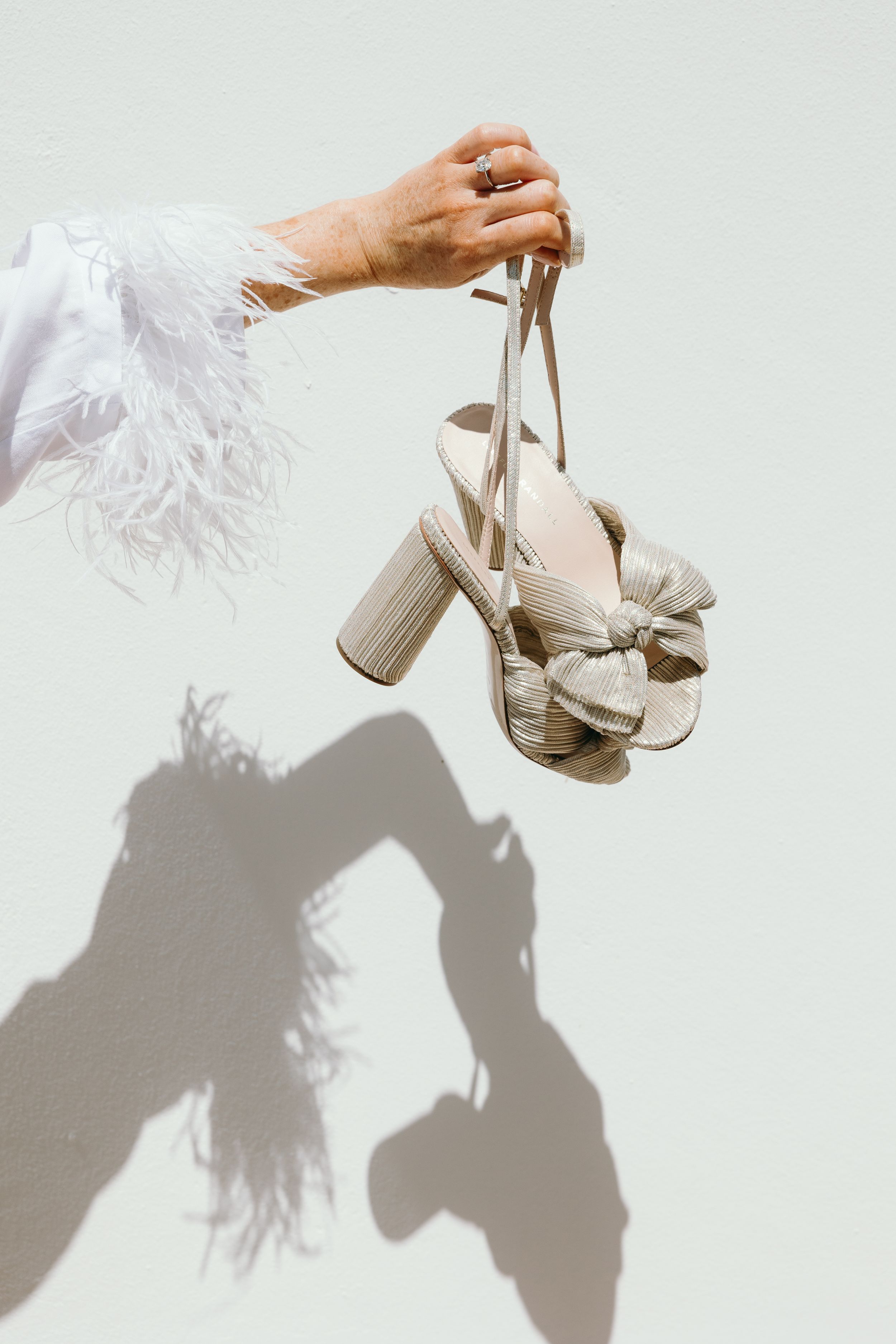 A hand holds elegant beige bow-adorned wedding shoes against a white wall casting dramatic shadows.