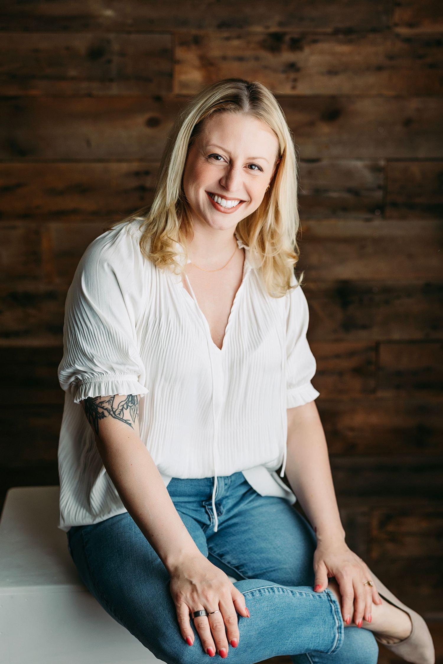Person wearing glasses and white tank top sits casually in light wash jeans against rustic wooden wall background.