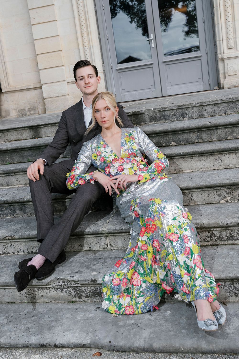 A couple sits on stone steps, with one person wearing a vibrant floral dress and the other in a dark suit.