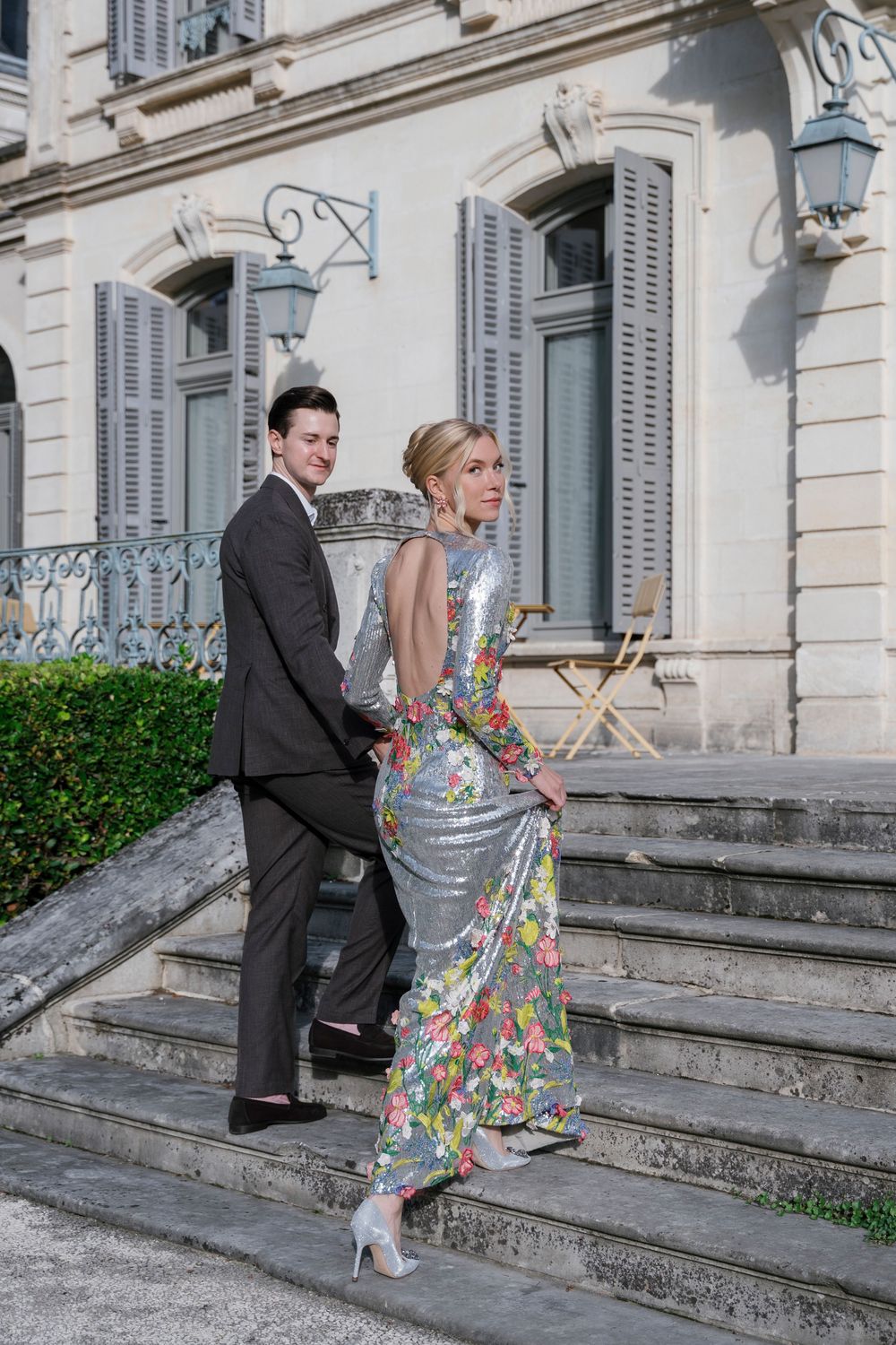A couple descends stone steps outside an elegant building with gray shuttered windows.