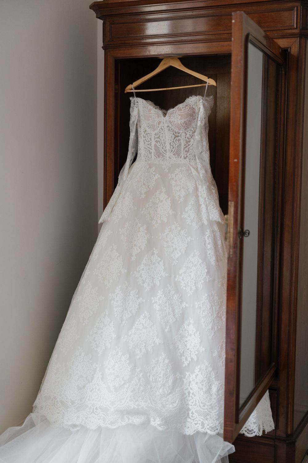 Elegant white lace wedding dress displayed on a wooden hanger against dark wood furniture.
